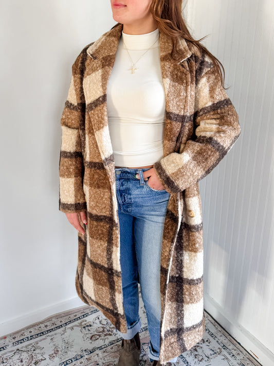 A person wearing the Nancy Coat, which is a long, plaid, button-down coat in beige, brown, and white colors, with side pockets and a slightly oversized fit.