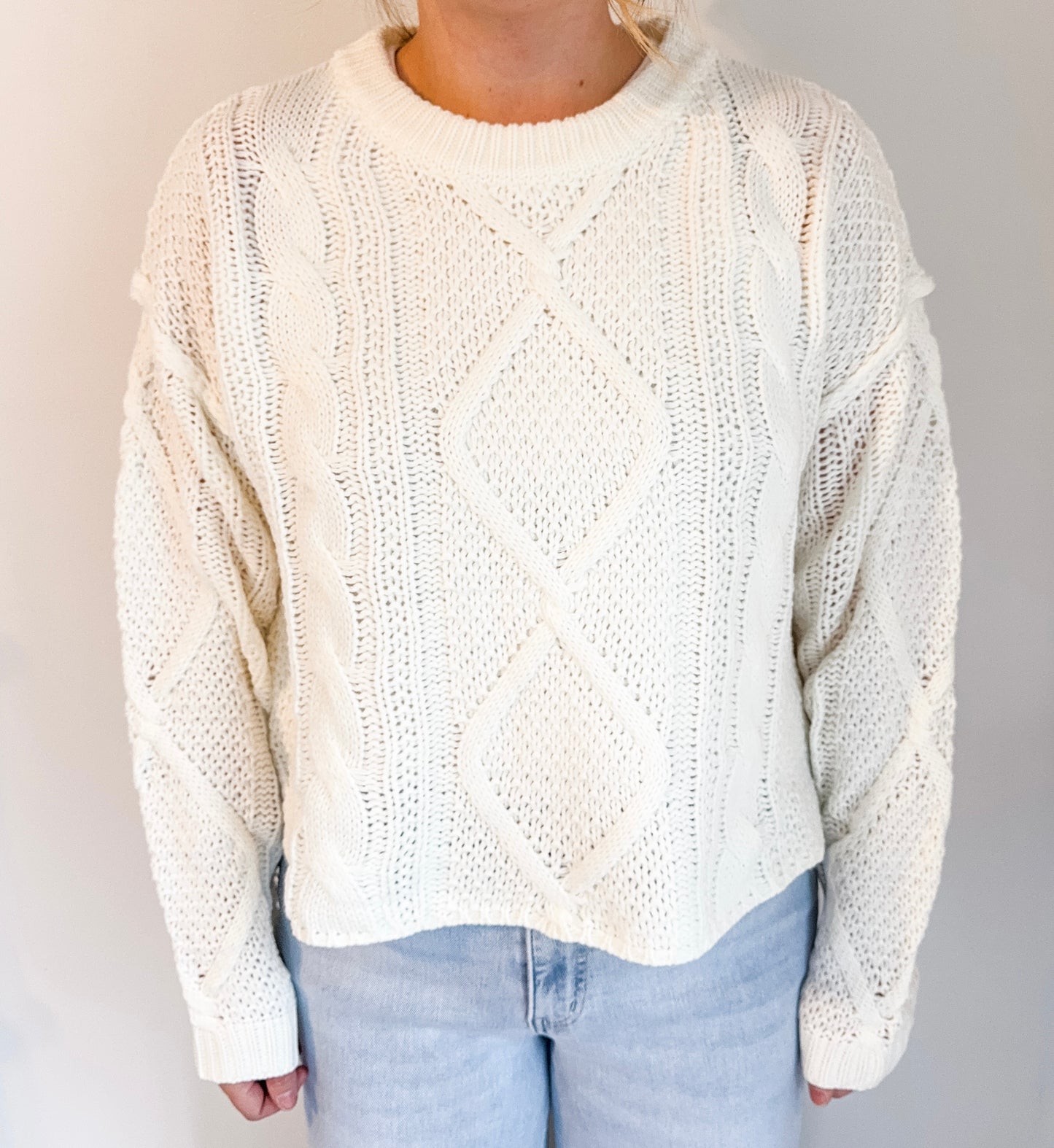Person wearing a cream knitted sweater and light blue jeans against a plain background